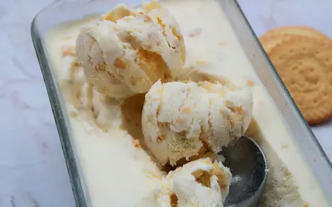Pyszne lody ciasteczkowe w szklanym naczyniu, z widocznymi kawałkami ciastek i gałkami lodów. Obok leży okrągłe ciastko.