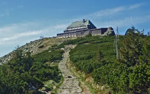Kamienista ścieżka prowadzi na szczyt, gdzie widać schronisko. Wokół bujna roślinność i skaliste zbocza.