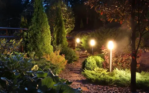 Oświetlenie bez prądu tworzy magiczną atmosferę w ogrodzie. Ścieżka z kamieni i drewna prowadzi przez bujną roślinność, oświetloną ciepłym blaskiem lamp.