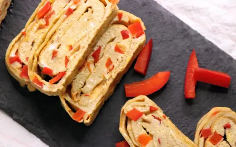 Roladki omletu tamago z kawałkami czerwonej papryki, ułożone na czarnej desce.