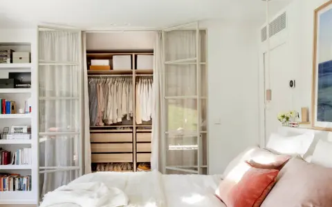 Dormitorio con vestidor integrado. Ropa organizada en armarios abiertos y estanterías. Cama con cojines rosas y manta blanca.