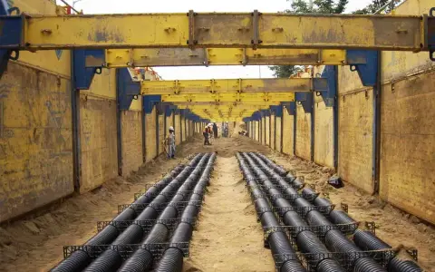 Trabajadores instalan tuberías en una zanja profunda, con estructuras metálicas amarillas para entibar zanjas y prevenir derrumbes.