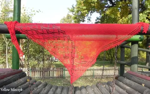 Czerwony, ażurowy szal, wykonany według darmowych wzorów z Ravelry, wisi na drewnianej konstrukcji placu zabaw.