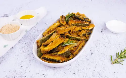 Zucchine gratinate al forno, dorate e croccanti, servite in un piatto ovale con rosmarino fresco. Accanto, ciotoline con pangrattato e olio.