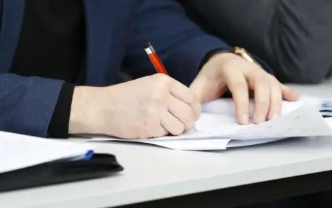 Ręka pisze długopisem po papierze, przygotowując przykładowy artykuł po angielsku na maturę.
