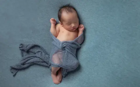 Neonato dorme avvolto in un telo blu, con le braccia sollevate e i pugni chiusi.