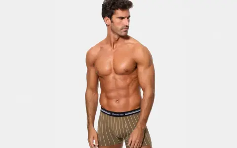 Hombre musculoso posando con calzoncillos de poliamida abriga, con rayas verticales blancas sobre fondo verde oliva.