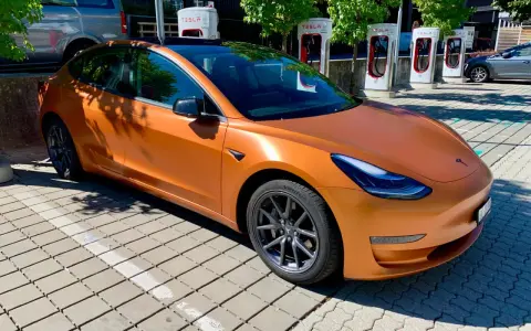 Pomarańczowa Tesla na tle ładowarek Supercharger.