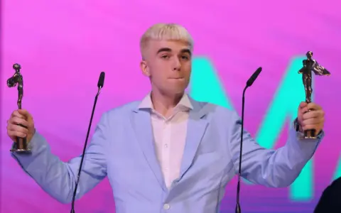 Mc Mata z dwoma statuetkami na scenie, w jasnoniebieskiej marynarce i białej koszuli.