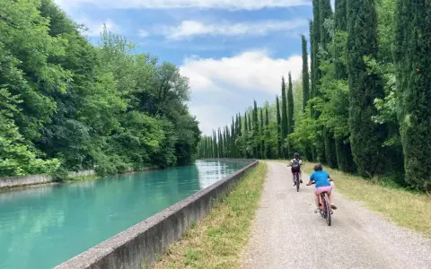 Bambini in bicicletta lungo la ciclovia del Mincio, con il canale turchese e filari di cipressi.