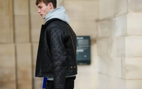 Uomo con un elegante outfit con giubbotto di pelle nero con monogrammi, felpa grigia e pantaloni scuri.