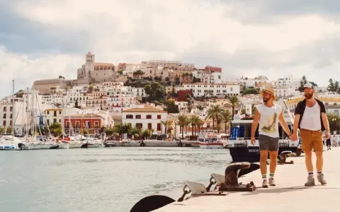 A gay couple walks hand-in-hand along a harbor in Spain, with a white-washed town and ancient fortress rising behind them.