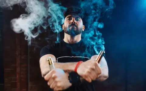 Hombre con barba y gorra, rodeado de vapor azul, sostiene dos vapers, uno dorado y otro cobre. La diferencia entre 0.8 y 1.2 ohm se siente en la calada.