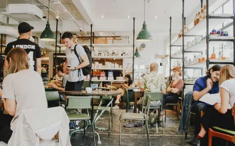 Gente en un café con mesas verdes y estanterías llenas de objetos. El **ruido restaurante** es palpable con las conversaciones y el movimiento.