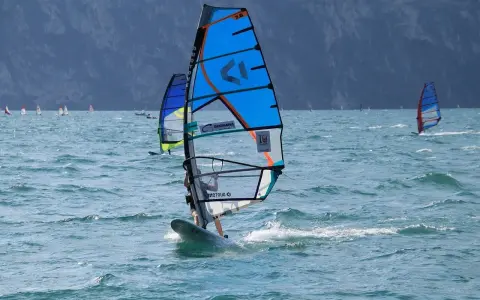 Osoba na desce windsurfingowej z niebieskim żaglem na wzburzonym morzu.