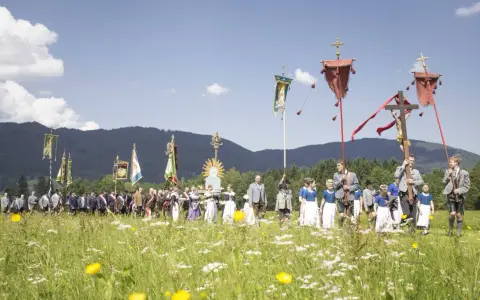 Menschen in Tracht und Uniformen nehmen an einer Prozession teil, die den Fronleichnam feiert. Fahnen und ein Kreuz werden getragen.
