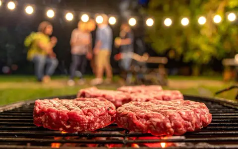 Frische Burger-Patties liegen auf dem Grill. Im Hintergrund feiern Freunde unter Lichterketten. So macht man Hackfleisch selber für den perfekten Grillabend.