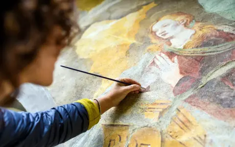 Une artiste restaure une fresque ancienne, sa main tenant un pinceau pour la préparation peinture. La scène montre une figure religieuse aux mains jointes.