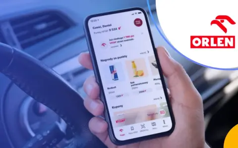Dłoń trzyma smartfon z aplikacją ORLEN Pay, pokazującą punkty VITAY i nagrody. Kierownica samochodu w tle.