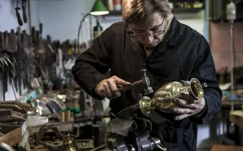 Un orfèvre au travail, martelant délicatement un vase en métal dans son atelier rempli d'outils.