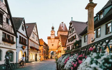 A charming street in gay Germany, with historic buildings, cobblestones, and flowers. Two people on bikes pause to take in the view.