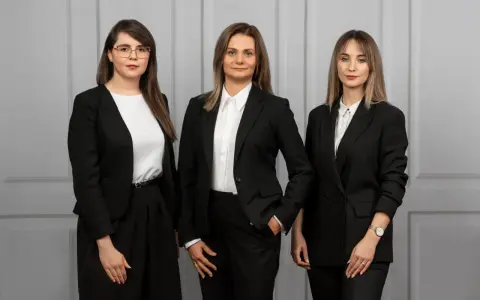 Trzy kobiety w czarnych garniturach stoją przed szarą ścianą.