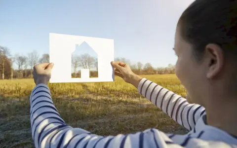 Szukasz idealnej działki pod dom 70m2? Kobieta trzyma wycięty z papieru dom na tle słonecznego pola i drzew.