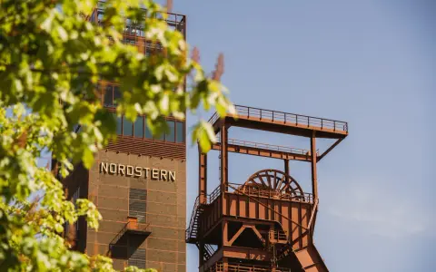 Industriearchitektur im Nordsternpark Gelsenkirchen: Ein markanter Förderturm und das Gebäude mit der Aufschrift "NORDSTERN" vor blauem Himmel.