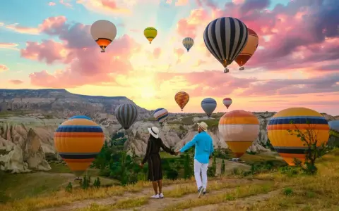 A couple holds hands, gazing at a sky filled with hot air balloons over the unique landscape of western Turkey at sunrise.