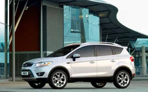 Biały Ford Kuga 2010 zaparkowany przed nowoczesnym budynkiem z przeszkloną fasadą.