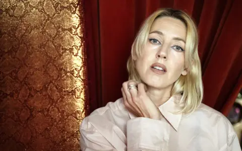 Veronica Maggio ung, med blont hår och blå eyeliner, poserar framför en gyllene ormskinnsmönstrad bakgrund och röda draperier.