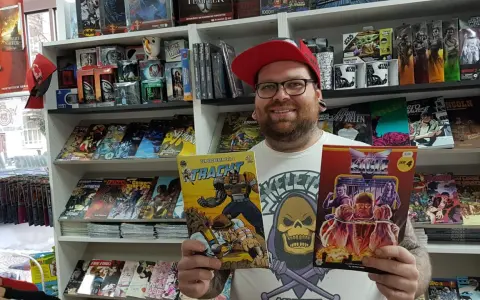 Mann mit Brille und roter Kappe hält zwei Comics in einem Comicladen.