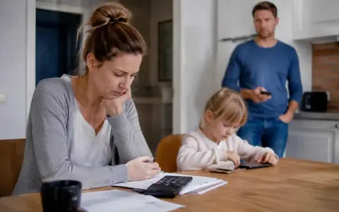 Zmartwiona kobieta z kalkulatorem przy biurku, dziecko pochłonięte telefonem, mężczyzna stoi z tyłu. Czy należy się opieka nad dzieckiem gdy żona nie pracuje?