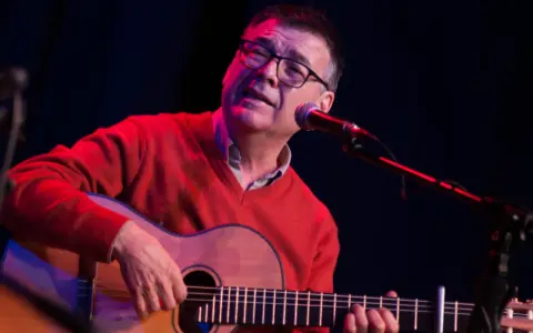 Mario Díaz, con gafas y un suéter rojo, toca la guitarra y canta frente a un micrófono en un escenario con luces tenues.