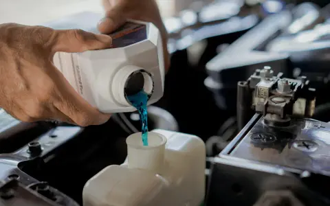 Hands pour blue coolant into a car's reservoir. This maintenance task is essential for preventing overheating, which can lead to costly car radiator replacement.