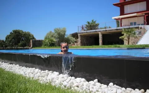 Niña sonriente en una piscina desbordante, con agua cayendo sobre piedras blancas y césped verde. Al fondo, una casa moderna y árboles frondosos bajo un cielo azul.