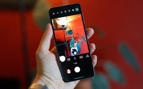 Dłoń trzyma smartfon z włączonym trybem PRO aparatu. Na ekranie widoczny jest kadr z rośliną w doniczce i lampą.