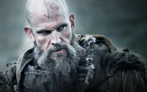 Uhtred z Bebbanburga, wojownik z tatuażami na głowie i długą, siwą brodą, spogląda groźnie.
