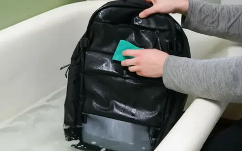 Se puede lavar una mochila en la lavadora. Alguien limpia una mochila negra en una bañera con agua y jabón.