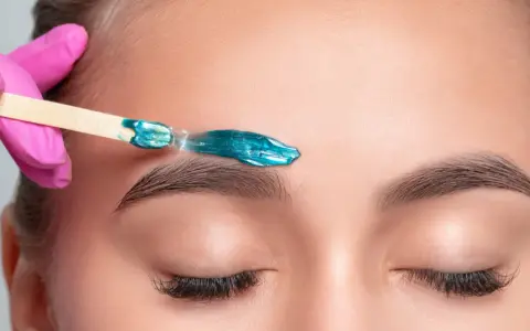 A pink-gloved hand applies turquoise wax to an eyebrow. This close-up shows the contrast between hot wax vs cold wax application for brow shaping.