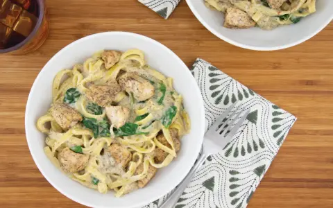 Delicioso plato de pasta alfredo con trozos de pollo y espinacas, servido en un tazón blanco sobre una tabla de madera.
