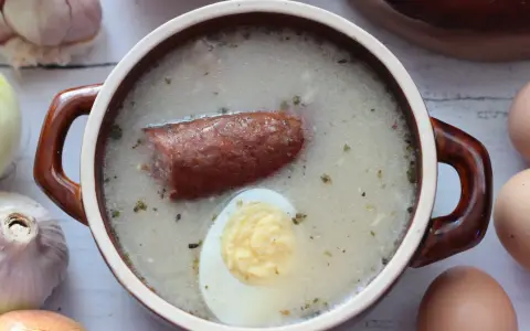 Aromaticzny zakwas na żurek w glinianym naczyniu, z kiełbasą i jajkiem. Obok czosnek, cebula i jajka.