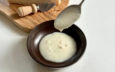 Una cucchiaiata di cremosa fonduta di parmigiano cade in una ciotola. Sul tagliere, un pezzo di Parmigiano Reggiano e un coltellino.