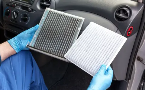 A dirty cabin air filter can make your car smell. A gloved hand holds a very dirty filter next to a clean, white one, showing the stark contrast.