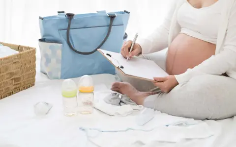Gravid kvinna skriver på en lista för packlista bb. Bredvid ligger en blå väska, korg med kläder, nappflaskor och bebiskläder.