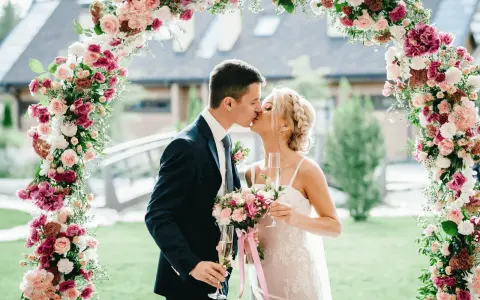 Novios besándose bajo un arco floral rosa y blanco, celebrando su boda barata con copas de champán.