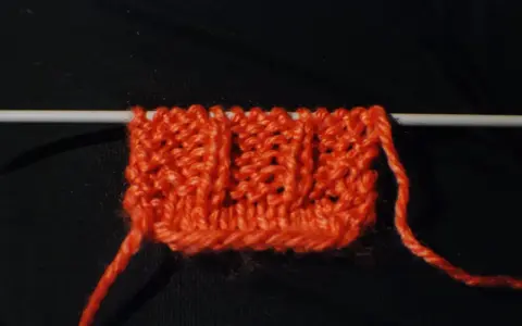 En orange stickad provlapp med ett mönster av vriden resår på en sticka. En lös tråd hänger ner.