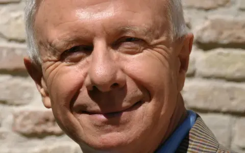Primo piano sorridente di padre Alberto Maggi, con rughe d'espressione e capelli grigi, sullo sfondo di un muro di mattoni.