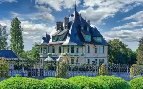 La villa Demoiselle, une demeure de caractère avec ses toits pointus et sa façade claire, se dresse fièrement derrière une grille ouvragée et des buissons taillés.