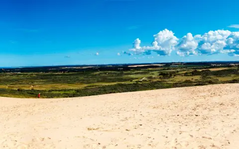 En person i röd jacka vandrar på en sanddyn vid Jyllands strand. Landskapet sträcker sig mot en blå himmel med vita moln.